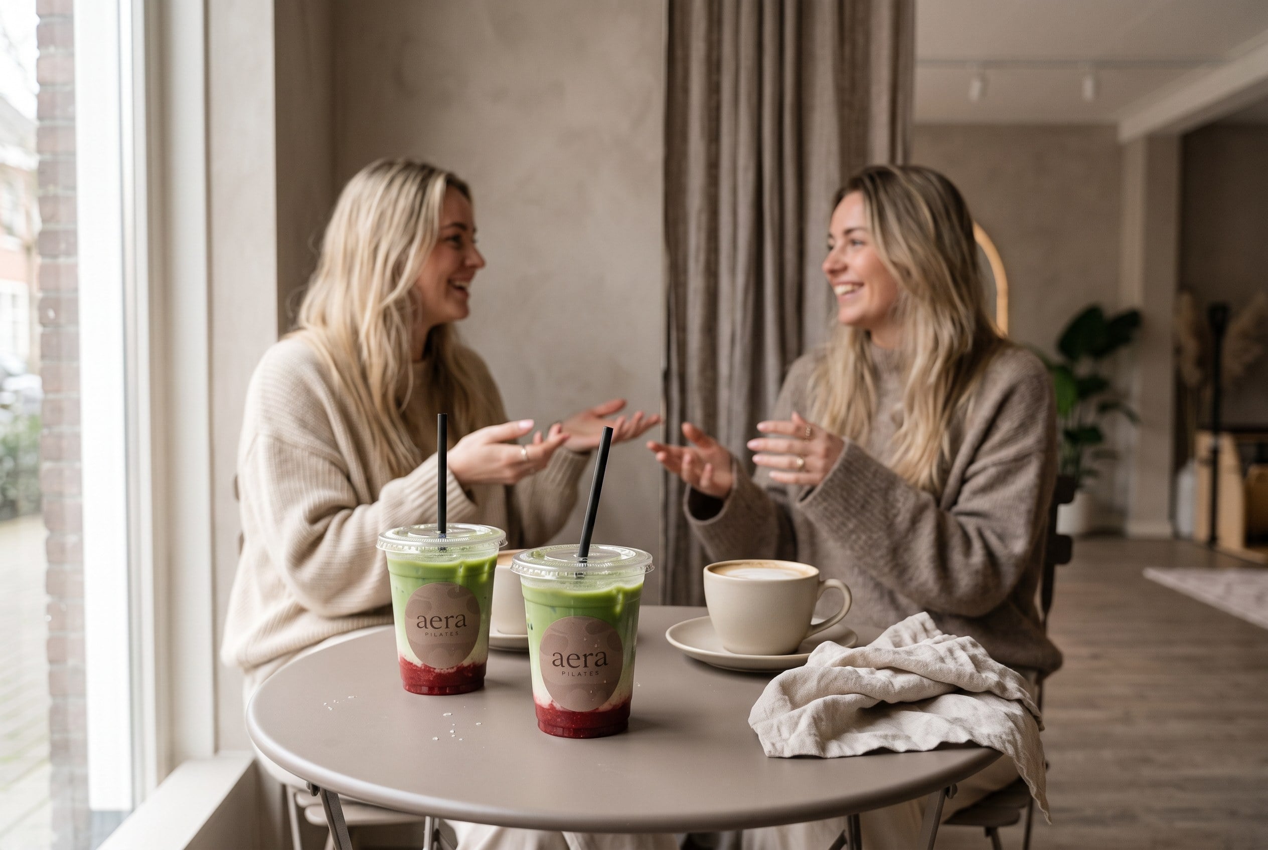 Two friends laughing at a bistro table with two aera strawberry matchas and a cappuccino